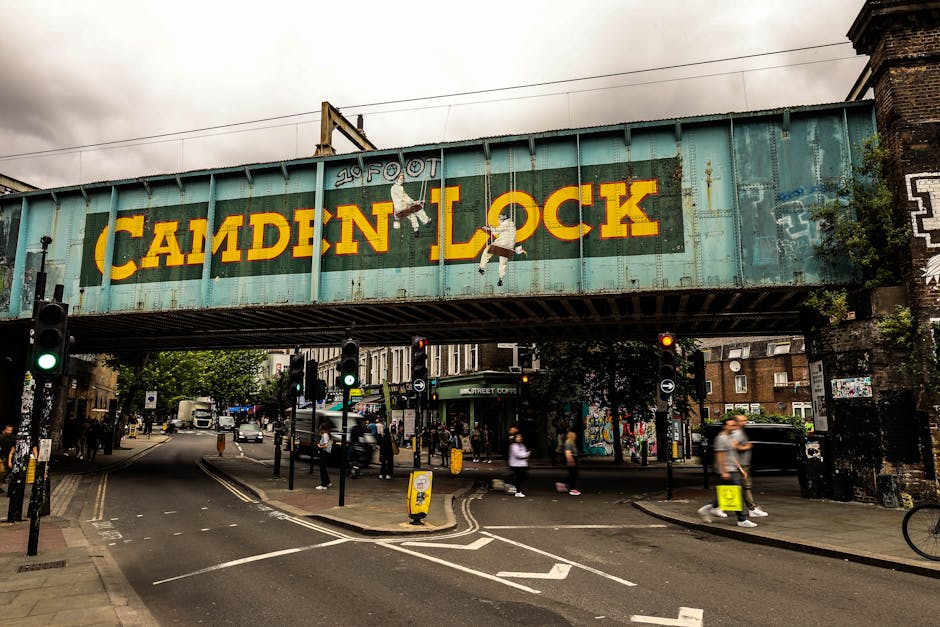 A large, weathered railway bridge spanning across a busy street in Lambeth, London, displaying a colorful mural with the text 'Camden Lock' in bold yellow letters. The bridge has a rusted, turquoise metal structure with visible rivets and supports. Painted figures of two children hanging from the letter 'L' add artistic detail. Below, several people are walking along the pavement, some pushing strollers or carrying bags, while others wait at multiple pedestrian crossings. Vehicles, including cars and delivery trucks, are seen moving through the intersection with traffic lights in green and red signals. The street is lined with brick buildings, lampposts, and street signage, capturing an urban scene associated with city life and dynamic movement, relevant to the logistics of home relocation and furniture transport in a typical Lambeth neighbourhood. Removals Lambeth may use such locations as part of their moving routes, coordinating the loading and unloading process near urban infrastructure. The environment is cloudy, with overcast skies providing diffuse natural light.