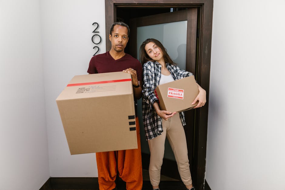 A man and a woman standing inside a doorway, each holding cardboard boxes for home relocation. The man, positioned on the left, has short dark hair and is wearing a maroon t-shirt and orange pants, holding a large cardboard box with a red tape seal. The woman, on the right, has long brown hair and is dressed in a checkered shirt over a white t-shirt and beige trousers, holding a smaller cardboard box labeled 'FRAGILE' with red tape. They are situated in a narrow corridor, with the door opening to the outside pavement visible behind them. The scene captures the loading process during a furniture transport or packing and moving operation, with the environment lit naturally and minimal equipment like the boxes and the doorway visible. This image is associated with house removals services offered by Removals Lambeth, supporting professional relocation activities in the context of packing, transportation, and furniture transport.