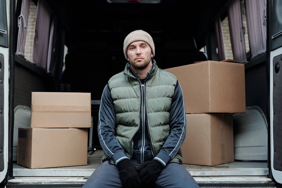 A man wearing a beige knit beanie, a green puffer vest over a navy hoodie with white stripe details, and black gloves, sits inside the open cargo area of a moving van. The van's interior is visible with several medium-sized cardboard boxes stacked neatly behind him. The boxes are sealed with packing tape, some are placed directly on the van's floor while others are stacked on top. The background of the image shows a driveway or pavement area, suggesting a home relocation setting. The lighting appears natural, indicating daytime. This scene captures the process of loading and organizing packed boxes for moving, exemplifying steps involved in packing and furniture transport, as highlighted by Lambeth removals services at removalslambeth.org.uk, focusing on efficient home moves and professional loading procedures.