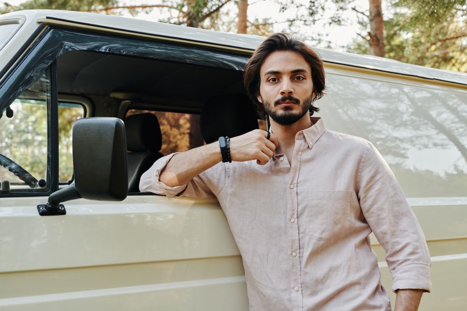 A man with shoulder-length dark hair and a beard, dressed in a beige button-up shirt with rolled-up sleeves, is leaning against the open driver's side door of a light-colored van. He holds a pair of sunglasses in his right hand, resting it near his neck, and looks directly at the camera with a neutral expression. The van is parked outdoors on a paved area, with trees and natural light filtering through the leaves in the background. Inside the van, visible through the open door, are black seats and part of a cargo space, suggesting it is used for home relocation or furniture transport. The scene reflects the process of packing and preparing for a house move, with the vehicle ready for loading or unloading furniture and packing boxes, which are not visible in this particular image. Removals Lambeth, a professional moving service provider, operates in a setting that emphasizes careful handling of belongings during home relocations.