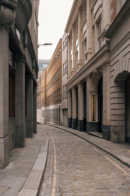 A narrow cobbled street in an urban area with tall, multi-storey buildings lining both sides. The buildings are constructed from stone and brick, featuring large windows and architectural details typical of city architecture. On the left side, a dark shopfront is visible, while the right side shows a building with an arched doorway and decorative facade elements. The street is empty of pedestrians and vehicles, with no visible signage or street furniture. Overhead, a single streetlight extends from the left side, illuminating the scene softly. In the context of house removals, the image depicts a typical access route for furniture transport or moving vans navigating narrow city streets, highlighting the importance of planning for tight street access during home relocation projects. This setting is relevant for London-area removals companies like Removals Lambeth, especially when coordinating moves in confined, historic urban environments like Brixton SW9.