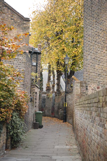 A narrow residential street with brick buildings on both sides, paved with uneven stone slabs, during autumn with fallen leaves scattered on the ground. There is a green wheelie bin placed against the left brick wall, and a tall lamppost with a black pole and a glass lantern is positioned along the street. The street is lined with trees that have yellow and orange foliage, some branches extending over the pathway. The scene is well-lit with natural daylight, and the surroundings suggest a quiet neighbourhood suitable for home relocation or furniture transport activities. Removals Lambeth could utilise such an area for loading or unloading during house removals in Lambeth, especially when navigating narrow street access for moving trucks.