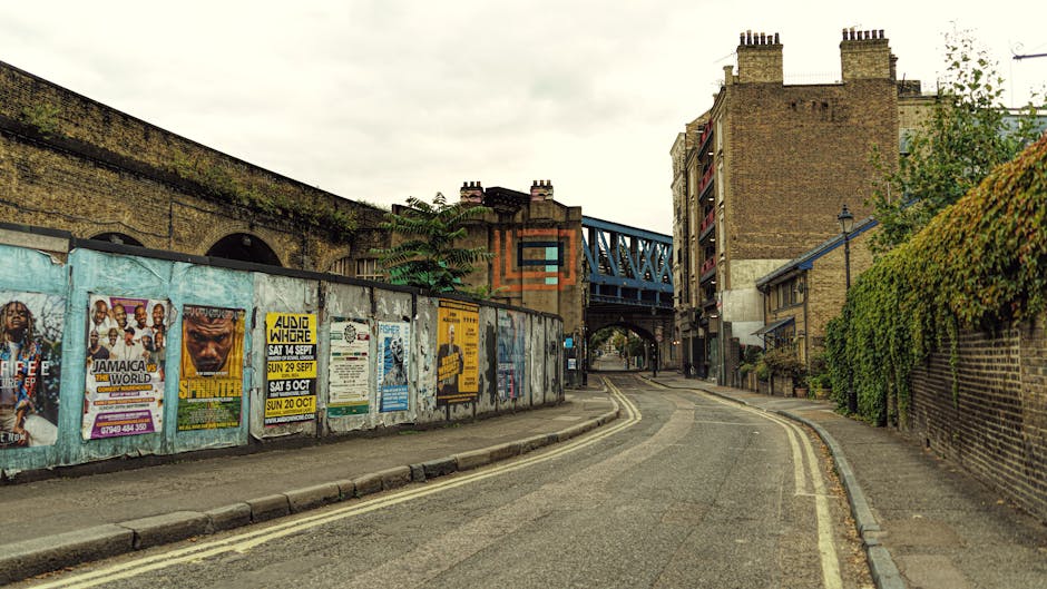 A narrow cobbled street in an urban area with tall, multi-storey buildings lining both sides. The buildings are constructed from stone and brick, featuring large windows and architectural details typical of city architecture. On the left side, a dark shopfront is visible, while the right side shows a building with an arched doorway and decorative facade elements. The street is empty of pedestrians and vehicles, with no visible signage or street furniture. Overhead, a single streetlight extends from the left side, illuminating the scene softly. In the context of house removals, the image depicts a typical access route for furniture transport or moving vans navigating narrow city streets, highlighting the importance of planning for tight street access during home relocation projects. This setting is relevant for London-area removals companies like Removals Lambeth, especially when coordinating moves in confined, historic urban environments like Brixton SW9.