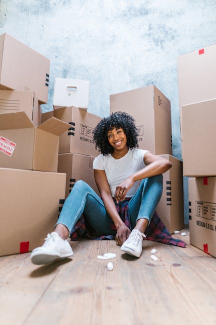 A young woman with curly black hair and a bright smile is sitting on a wooden floor inside a room surrounded by stacked cardboard boxes of various sizes, some with red and black labels indicating frag