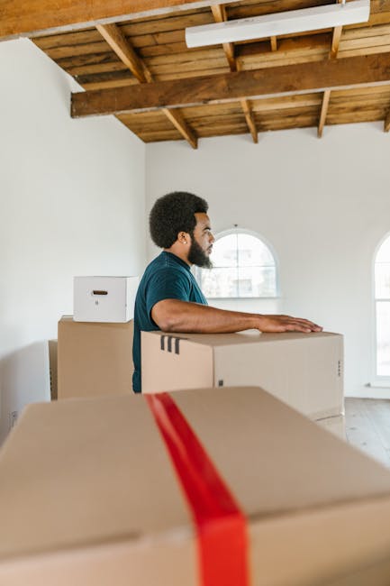 A man with a beard and curly hair, dressed in a teal shirt, stands inside a bright, spacious room with a high wooden ceiling supported by exposed beams and white walls. He is positioned behind several cardboard boxes, some sealed with red packing tape, which are stacked on the floor. The boxes vary in size and are likely used for packing household items during a home relocation, as part of furniture transport and moving services. The room has two large, arched windows allowing natural light to illuminate the space, and the man appears to be preparing for or engaged in a loading process. The environment suggests an interior in the midst of packing and moving preparations, typical of professional removals provided by Removals Lambeth, supporting detailed and organized house removals in the Clapham Common area.