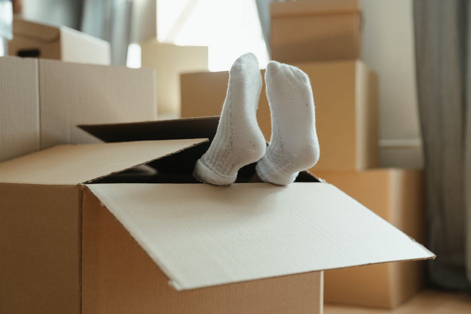 A pair of white socks are positioned upright inside a cardboard box, which is partially open, revealing the interior. Surrounding the box are several other packed cardboard boxes of varying sizes, some stacked in the background near the window, where daylight filters into the room. The scene appears to be inside a home during a house move, with boxes prepared for packing or unpacking. The environment is well-lit with natural light, and the boxes are made of brown corrugated cardboard, indicating the packing process involved protective packaging materials. This setup suggests a home relocation or moving process typical of furniture transport and packing activities, with the involvement of professional removal services such as Removals Lambeth, who handle logistics and loading for house removals.