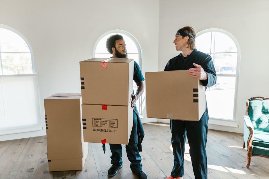 A man with a beard and curly hair, dressed in a teal shirt, stands inside a bright, spacious room with a high wooden ceiling supported by exposed beams and white walls. He is positioned behind several cardboard boxes, some sealed with red packing tape, which are stacked on the floor. The boxes vary in size and are likely used for packing household items during a home relocation, as part of furniture transport and moving services. The room has two large, arched windows allowing natural light to illuminate the space, and the man appears to be preparing for or engaged in a loading process. The environment suggests an interior in the midst of packing and moving preparations, typical of professional removals provided by Removals Lambeth, supporting detailed and organized house removals in the Clapham Common area.
