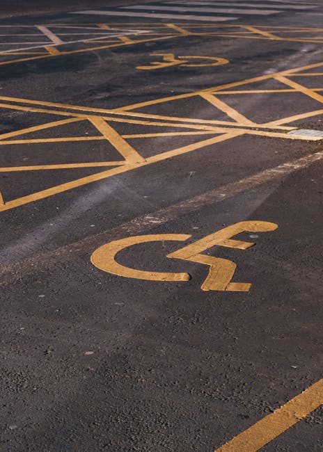 A close-up view of an asphalt parking lot featuring multiple disabled parking spaces marked with yellow painted lines and wheelchair symbols. The spaces are designated with bold yellow lines and symbols, with some spaces positioned side by side and others at slight angles, all clearly visible and well-defined. The parking lot surface appears smooth but has some minor wear and patching. There are no vehicles or other objects inside the marked spaces, which are located adjacent to the building entrance. The image captures the area where furniture, boxes, or moving equipment from a home relocation might be loaded into a nearby van or truck. The scene reflects a typical exterior setting for coordinating moving logistics in Lambeth, where Removals Lambeth provides professional house removal services, including parking arrangements and loading operations.