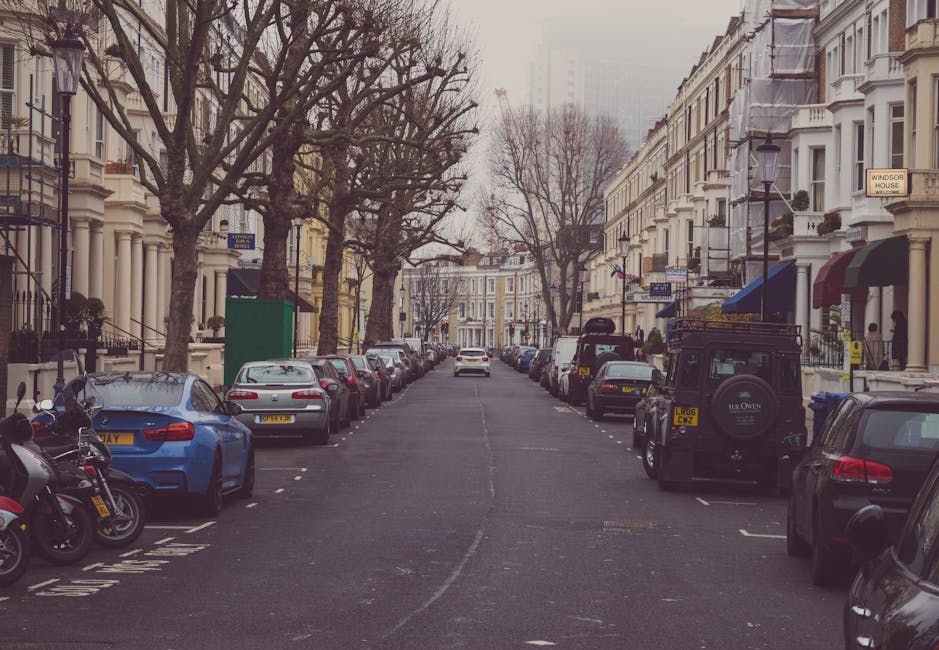 A residential street in Lambeth lined with multi-storey Victorian-style terraced houses featuring white facades, bay windows, and decorative railings. Several leafless trees are planted along the pavement, which is occupied by parked cars and motorcycles on both sides of the street. A black Land Rover Defender with a roof rack and spare tire mounted on the rear is parked near the sidewalk, close to the entrance of the property, which is being used during a home relocation process. Large cardboard boxes, wrapped furniture, and packing materials are visible near the entrance, indicating ongoing packing or loading activities. The scene shows a loading area where movers are likely arranging furniture and boxes to load into a van, although the vehicle itself is not visible. Soft natural light illuminates the scene, and there are several signs and street lamps along the pavement. The setting illustrates typical elements involved in a house removal or furniture transport in Lambeth, consistent with the services offered by Removals Lambeth.