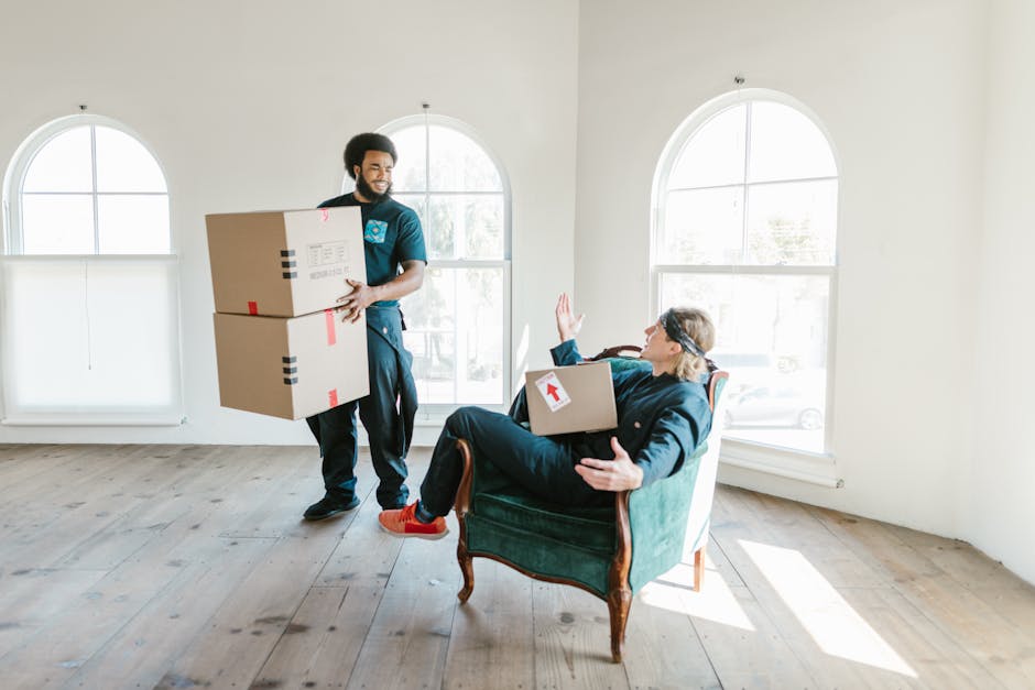 A professional removals worker from Removals Lambeth is inside a bright room with white walls and two large, arched windows, lifting two medium-sized cardboard boxes with red tape and labels. The worker, dressed in dark clothing, is smiling and carrying the boxes towards a seated elderly woman. The woman, wearing glasses and a dark jacket, is seated in a vintage-style armchair with wood accents, holding a cardboard box with a red arrow sticker, while raising her hand to gesture or wave. The room has a light wooden floor and minimal decoration, and natural daylight is streaming through the windows, illuminating the moving process involved in house relocation or furniture transport. This scene captures the careful handling and packing involved in home moves, with the involvement of professional services to facilitate the relocation process.