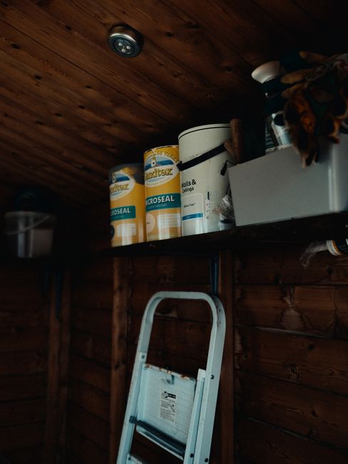 Inside a small, low-ceiling loft space with wooden wall paneling and a wooden ceiling, a dark-colored ceiling spotlight is visible. On a narrow wooden shelf mounted on the wall, there are three large cylindrical containers of paint or sealant, labeled 'Durostick' and 'Microseal,' with one beige container and two white and yellow ones. To the right of the containers, an open white box with various miscellaneous items, possibly tools or packing materials, is present. Below the shelf, a white folding step ladder leans against the wooden wall, indicating recent or upcoming home relocation activities involving furniture transport and packing in confined spaces. The environment suggests a storage or utility area within a home undergoing a move, with visible materials and equipment used in the moving process. Removals Lambeth could support such relocation tasks, especially where access solutions are needed for spaces with low ceilings.