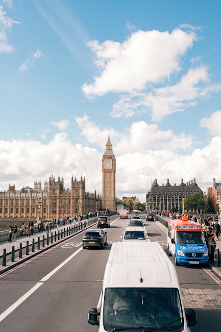 A busy urban street scene in London with several white and dark-colored vans, likely used for house removal and furniture transport, parked along the pavement. In the background, the iconic Big Ben clock tower and the Palace of Westminster are visible under a partly cloudy sky, indicating daytime. The street is lined with a black metal railing separating the road from the sidewalk, where pedestrians are walking. Vehicles are moving along the road, and a van is positioned near the curb, possibly in the process of loading or unloading. The overall environment suggests a typical city setting suitable for home relocation logistics, with clear weather providing good visibility for the loading and transport process. This scene exemplifies the operational context of removals services, as provided by companies like Removals Lambeth, involved in packing, moving, and furniture transport within the city.