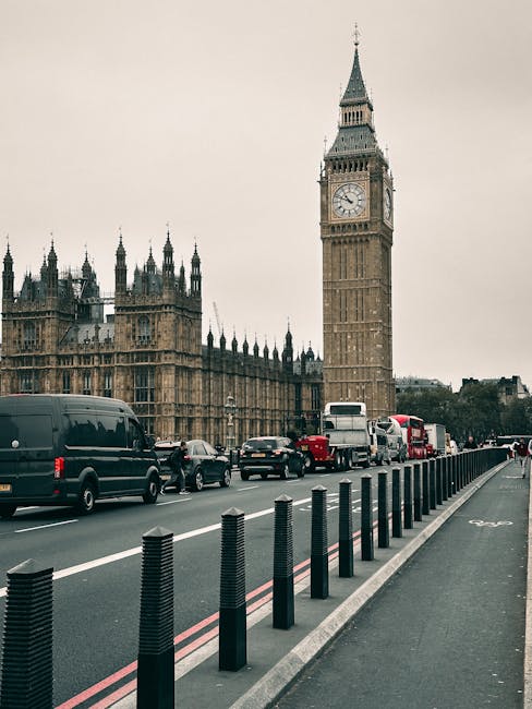 A city street near the Palace of Westminster in London, with the clock tower known as Big Ben prominently visible in the background. The street features a row of black bollards along the pavement, with vehicles including vans, cars, and double-decker buses queued in traffic. The scene appears to be during daytime with overcast skies. In the foreground, there is a loading zone adjacent to the pavement, likely used for furniture transport or home relocation activities. Removals Lambeth occasionally undertakes house moving services, and this busy urban environment reflects the logistical considerations involved in packing, loading, and transport during a same day move or home relocation process in central London.