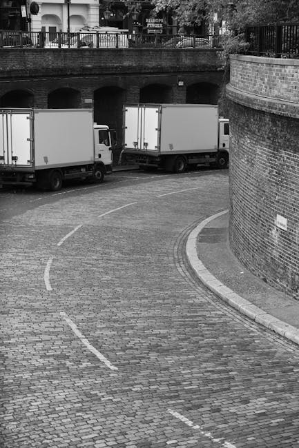 A busy urban street scene in London with several white and dark-colored vans, likely used for house removal and furniture transport, parked along the pavement. In the background, the iconic Big Ben clock tower and the Palace of Westminster are visible under a partly cloudy sky, indicating daytime. The street is lined with a black metal railing separating the road from the sidewalk, where pedestrians are walking. Vehicles are moving along the road, and a van is positioned near the curb, possibly in the process of loading or unloading. The overall environment suggests a typical city setting suitable for home relocation logistics, with clear weather providing good visibility for the loading and transport process. This scene exemplifies the operational context of removals services, as provided by companies like Removals Lambeth, involved in packing, moving, and furniture transport within the city.