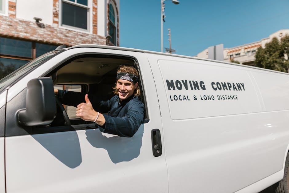 A smiling male mover with long hair, wearing a dark bandana and casual clothing, is sitting in the driver’s seat of a white moving van labeled 'MOVING COMPANY LOCAL & LONG DISTANCE'. He is giving a thumbs-up gesture through the open window. The van is parked in an urban setting with buildings, a streetlight, and a clear blue sky visible in the background. The scene suggests the initial stages of a home relocation, with the vehicle prepared for furniture transport, packing, and loading activities as part of the moving process handled by Removals Lambeth. Inside the van, there are visible cardboard boxes and protective blankets, indicating ongoing packing and loading operations for a house move, consistent with services offered for house removals and furniture transport in the Lambeth area.
